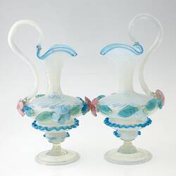 Pair of blue blown-glass ewer, spiral fluted neck, serpentine handle, flattened bulbous body layered with pink, blue, green flowers and leaves.