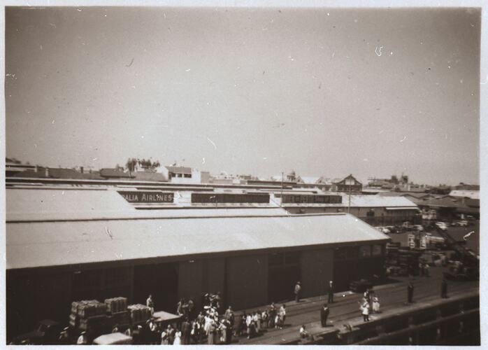 View of wharves from a ship. Long shed-like building with people in front on the dock.