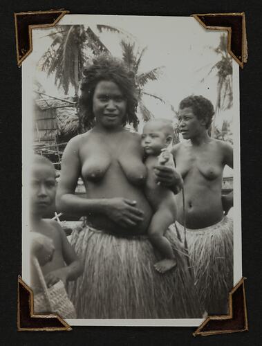 Two women wearing grass skirts, one woman is holding a baby with child at side.
