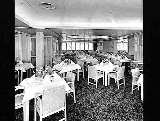 Ship interior. Dining room with chairs and tables. Tablecloths and flower vases decorate each table.