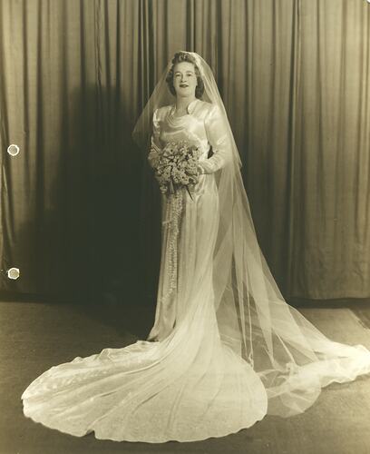 Bride in wedding dress with train and veil. Holding floral bouquet.