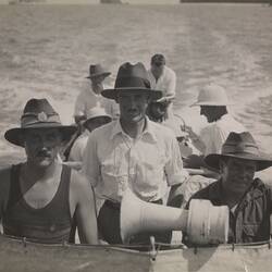 Seven men on a vessel, three posing. Water behind them. Six wear hats. Loud speaker in front of them.