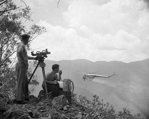 Two men with a tripod and radio watch a helicopter spray in a mountainous setting.