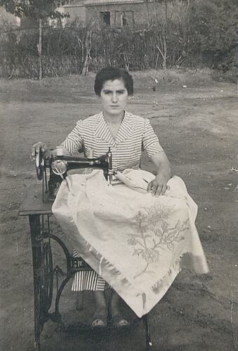 Woman sits at sewing machine set up outdoors. She is hemming a blanket.