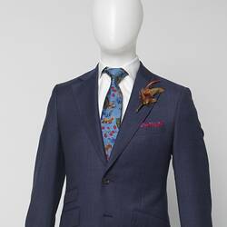 Detail of blue suit jacket with thin red square check pattern. White shirt, blue patterned tie. Brown feather corsage, red patterned kerchief strip at pocket.