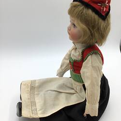 Profile, seated doll with red, green, white top. White apron over black skirt. Red hat, brown hair.