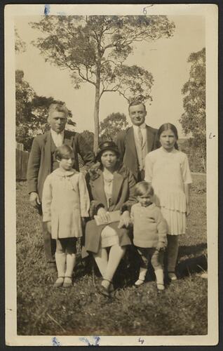 Two men stand behind seated woman. Three children stand beside her. Trees in background.