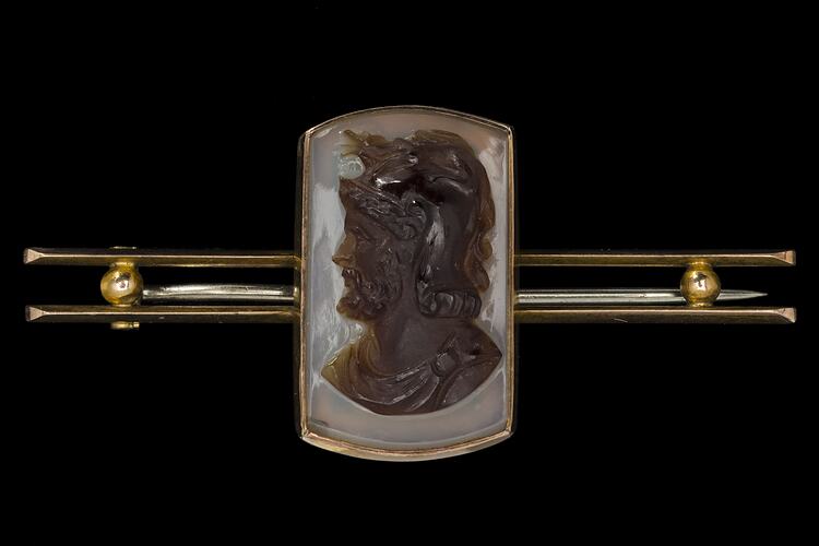 Gold bar brooch with centrally placed, rectangular cameo. Made of dark brown hardstone the cameo features a bearded male bust facing left with lighter brown background.