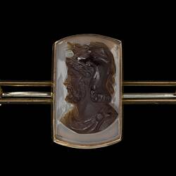 Gold bar brooch with centrally placed, rectangular cameo. Made of dark brown hardstone the cameo features a bearded male bust facing left with lighter brown background.