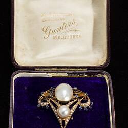 Delicate gold inverted triangle brooch with two main pearls framed by seed pearls and blue paste. A fine gold chain and pin extend from top left corner. In box.