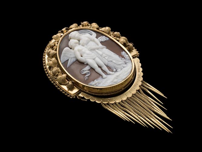 Oval cameo brooch with white winged figure and female, brown background. Ornate gold border and hanging fringe.