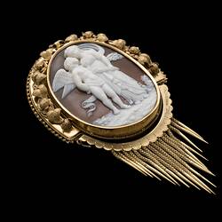 Oval cameo brooch with white winged figure and female, brown background. Ornate gold border and hanging fringe.