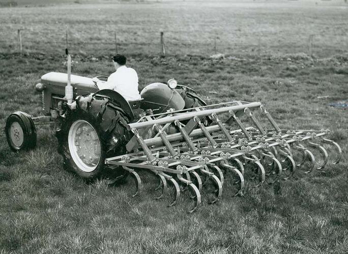 Rear view of a man driving a tractor coupled to a spring tine cultivator.