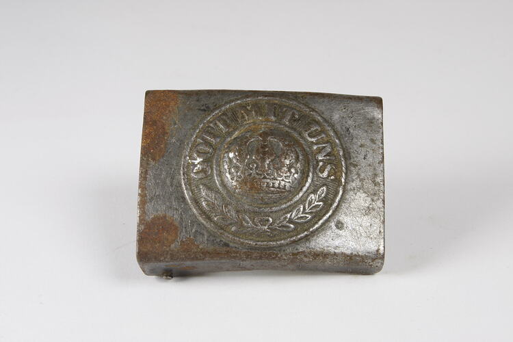 Silver metal belt buckle with crown and inscription in centre, patches of rust and scratches across surface.