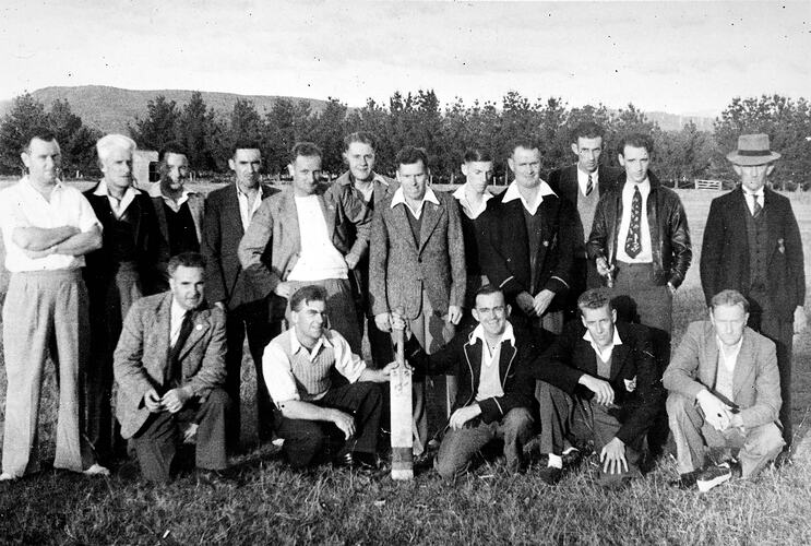 Two rows of men wearing regular clothes pose with a cricket bat.
