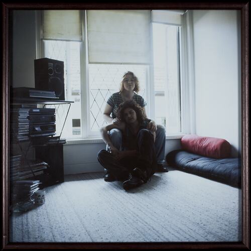 Two women sit propped up against window sill. Sound system at left, rolled mattress at right.