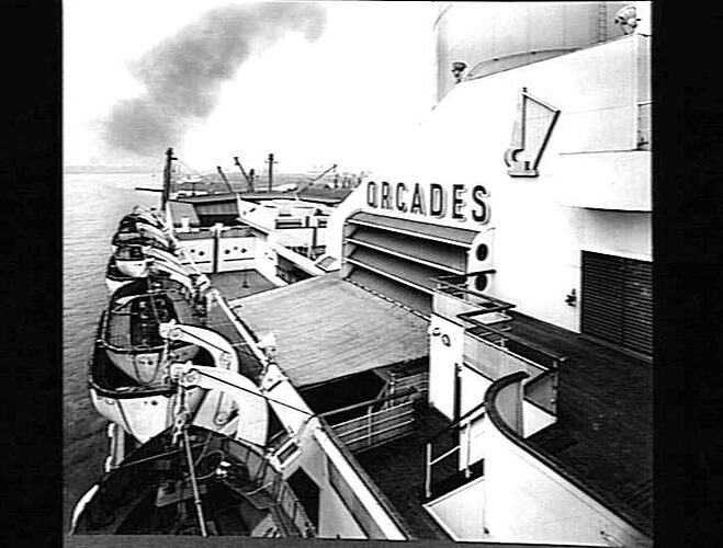 Onboard ship deck. Life boats at left. Ship name, Orcades, on right.