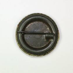 Reverse of round metal badge with pin fastener.