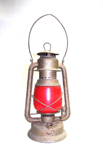 Portable red oil lantern with metal frame and wire handle.