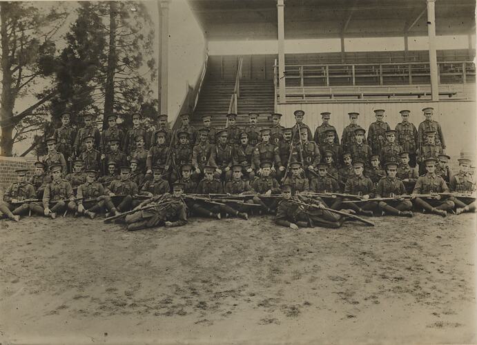 Group portrait of men in military uniform arranged in four rows with two men laying on ground in front.