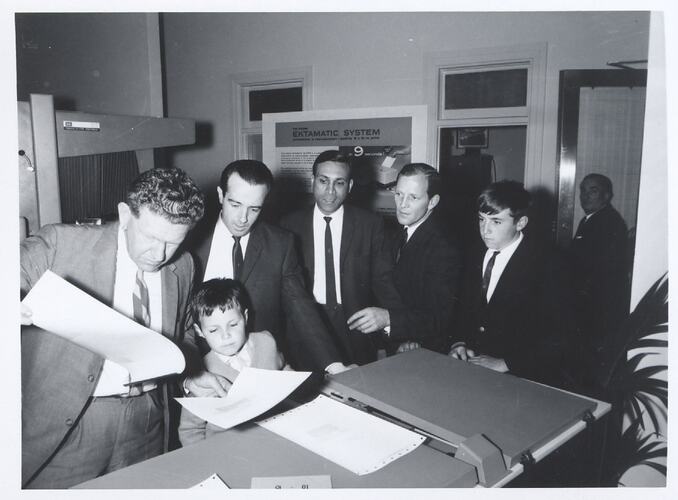 Men and boys in suits inspecting paper sheets.