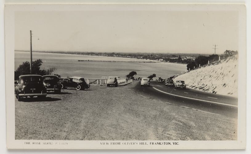 View of beach from hill with road and parked cars.