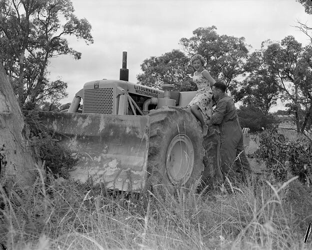 Man helping a woman on to a tractor with a front end plow in bushland.
