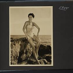 Album page with photograph, black and white, woman posing in polka dot bikini, leaning against rocks.