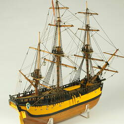 Three quarter view of model ship with wooden hull painted yellow and three masts.