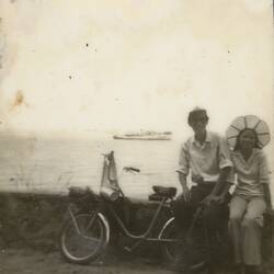 Man and woman sitting on a wall overlooking water with a bicycle to their right. Ship in background.