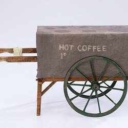 Miniature wooden coffee hand cart with two legs, two wheels. Fabric cover on top.