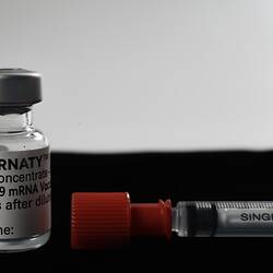 Glass vial with white printed label and metal cap beside syringe with black text and red cap.