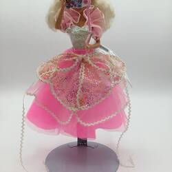 Barbie doll with layered pink and white dress. She holds a fancy dress mask.