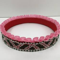 Round beaded crown with pink ruffled ribbon trim around the top. Green and white plastic beads.