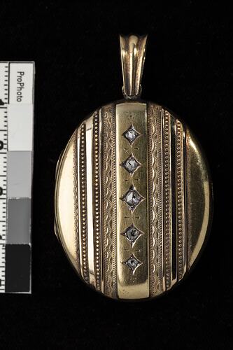 Gold oval locket with vertical row of diamonds framed by varied ornate vertical lines. Loop at top.