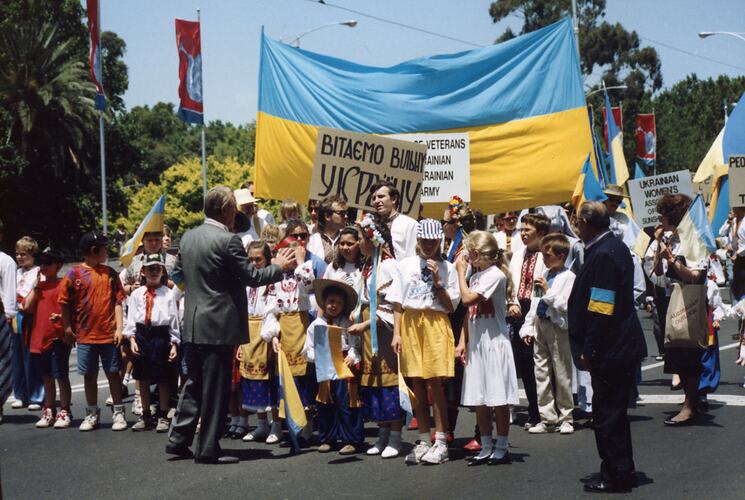 Group marching on street at public rally. Some wear yellow and blue and they have a large yellow/blue Ukrainian flag behind them.