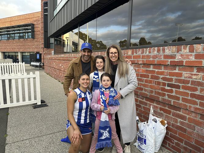 Man, woman and two girls pose with kneeling female footballer wearing blue and white striped football jumper. Building behind them.