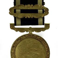 Round gold coloured medal with engraved text, black and gold ribbon attached.
