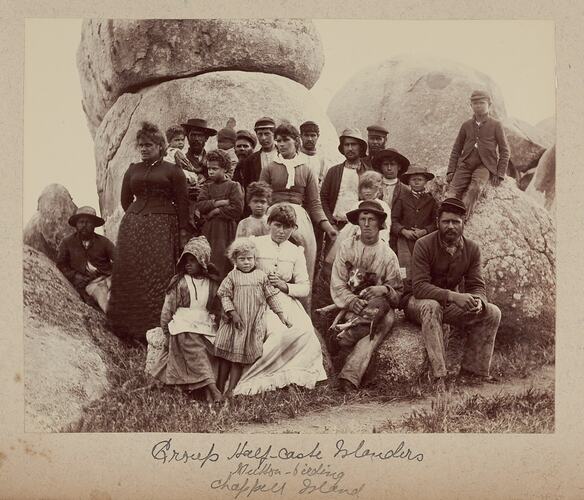 Men, women and children in front of granite boulders. One man holds a dog on his lap.