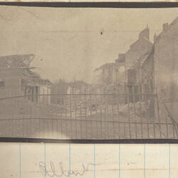 Street with damaged buildings and piles of debris, iron fence in foreground.