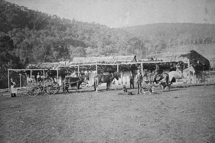 Bark roofed buildings. Carriage drawn by four horses at left and two men on horseback at right.