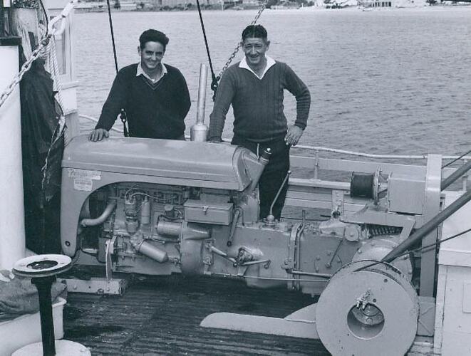 Two men looking at a tractor engine and winch, which has been mounted on a boat.