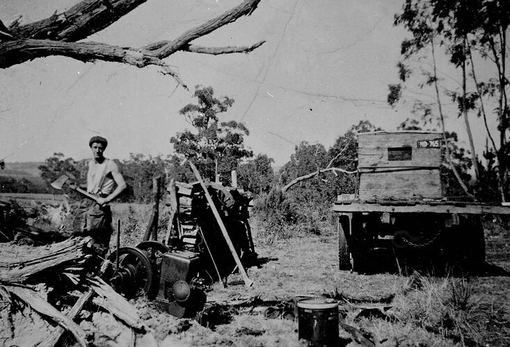 Man chopping wood with axe. Machinery and truck at right.