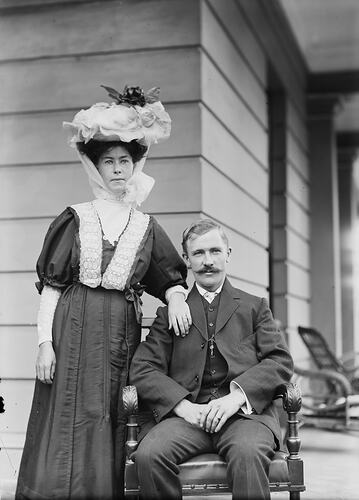 Seated man with woman standing beside him on a veranda. Her arm rests on his shoulder.