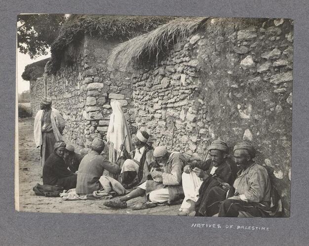 Villagers sitting against the drystone wall of a building with a thatched roof.