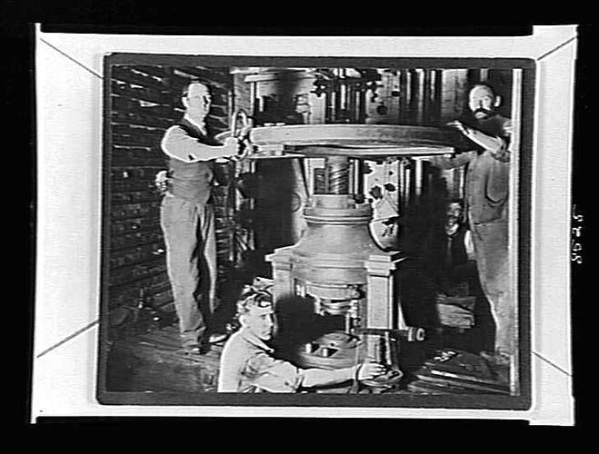 Four men around a large metal press in a small wooden room.