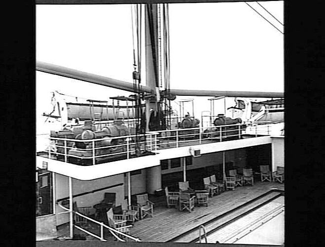 Ship deck with winch platform overlooking empty rectangular swimming pool and chairs.
