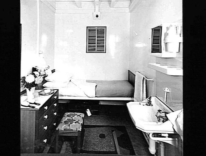 Ship interior. Single bed against wall. Chest of drawers at left, handbasin at right.