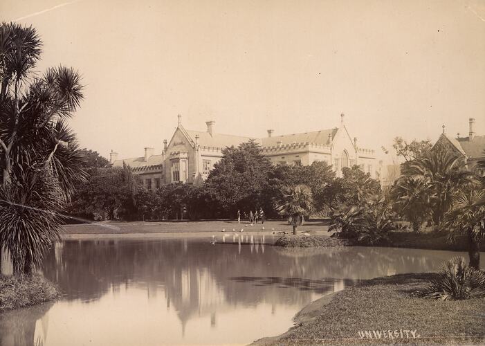 Buildings surrounded by a formal garden, which also contains an ornamental lake. Male students, wearing academic gowns, are near the lake watching ducks feed.