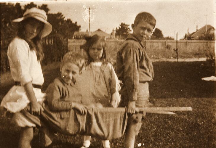 Boy & girl carrying stretcher, with another boy sitting in it and another girl standing beside, in backyard.